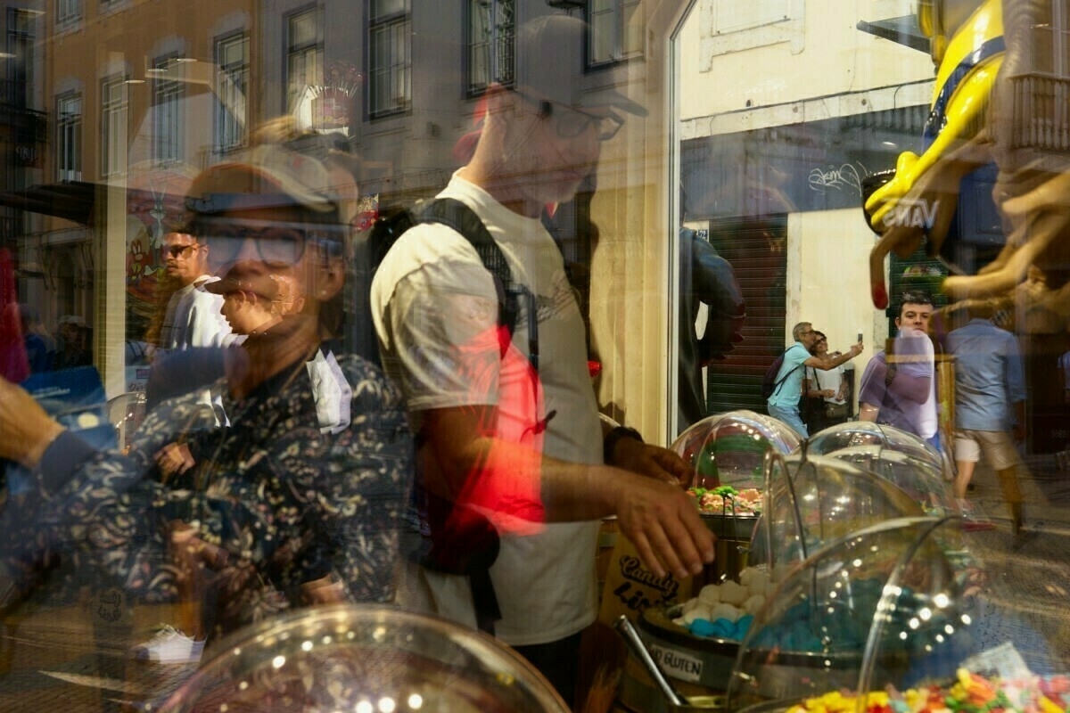 People are reflected in a shop window displaying various sweets and candies, creating a layered and busy scene.