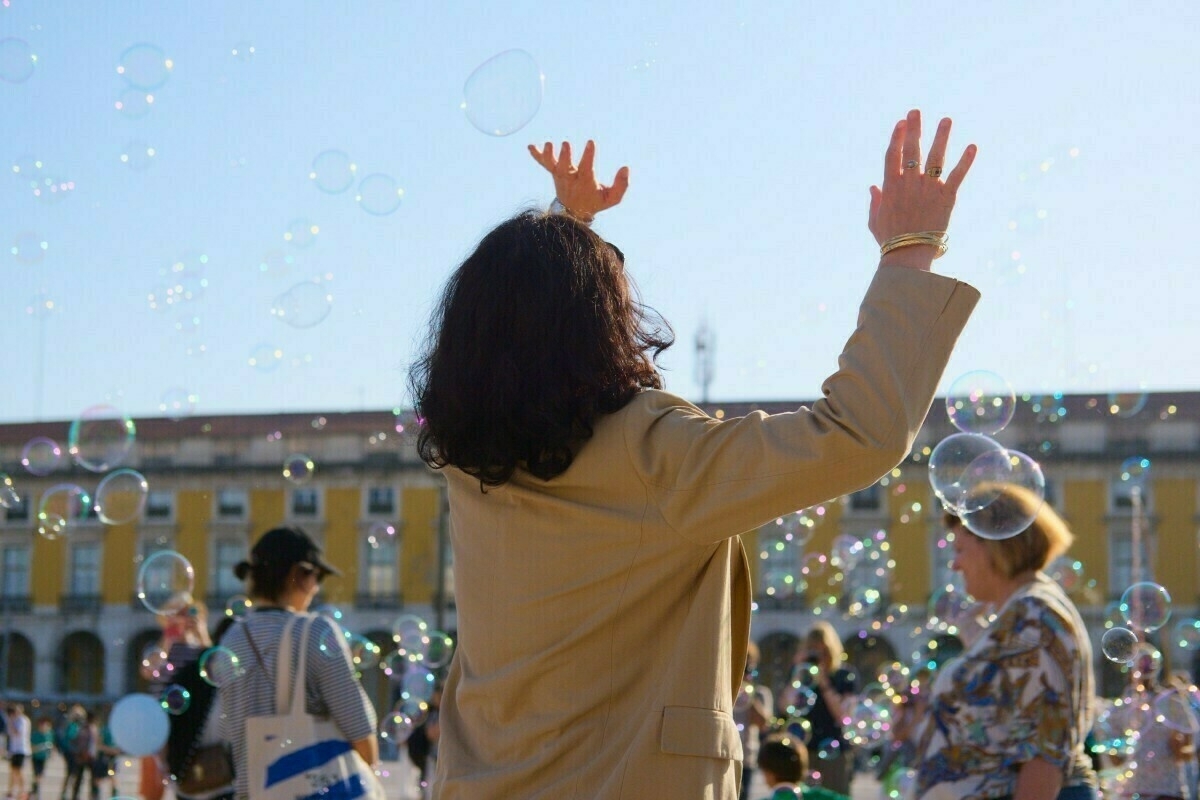 A person with long hair reaches upward towards bubbles floating in the air, surrounded by people in an outdoor setting with historic-looking buildings in the background.