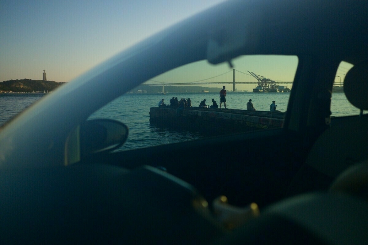 A serene waterfront scene is viewed through a car window, featuring people sitting on a pier with a bridge and cranes in the background.