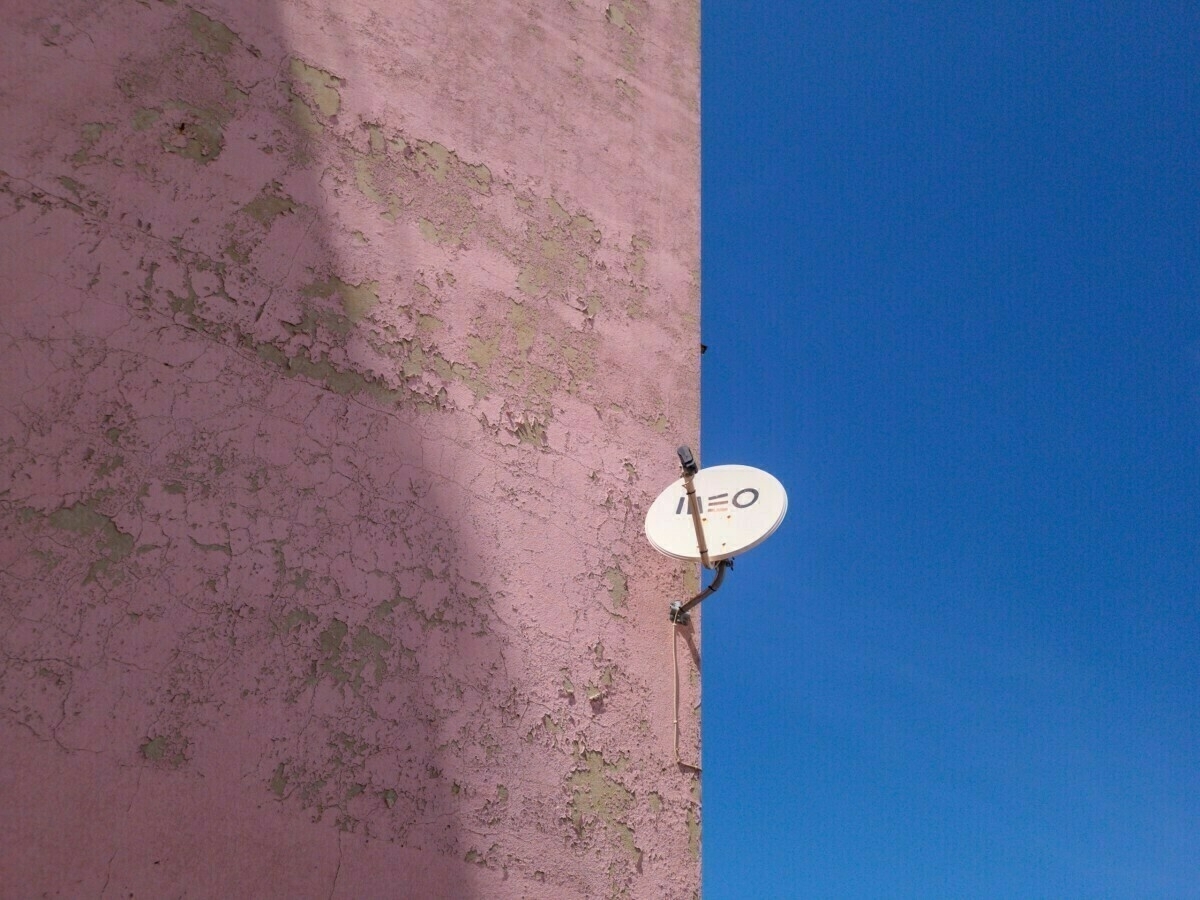 A satellite dish is mounted on a weathered, pinkish wall against a clear blue sky.