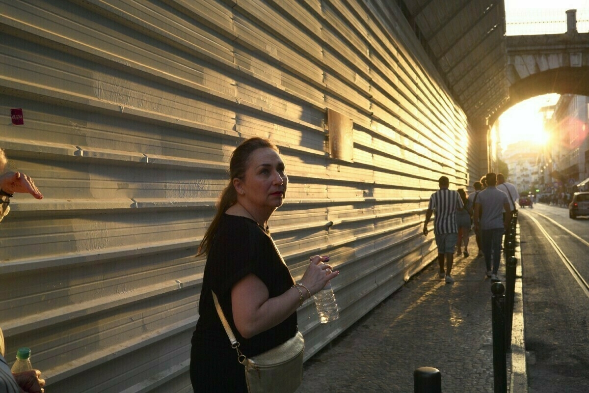 A woman holding a water bottle walks along a sunlit street with several other people in the background.