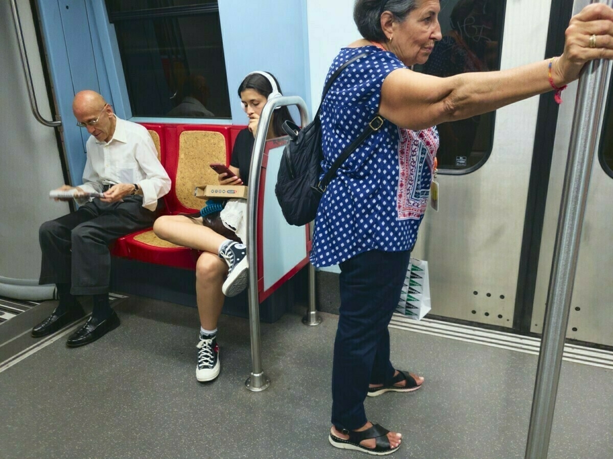 A young person and an older man are seated on a train, both focused on their phones, while a woman stands holding onto a pole.