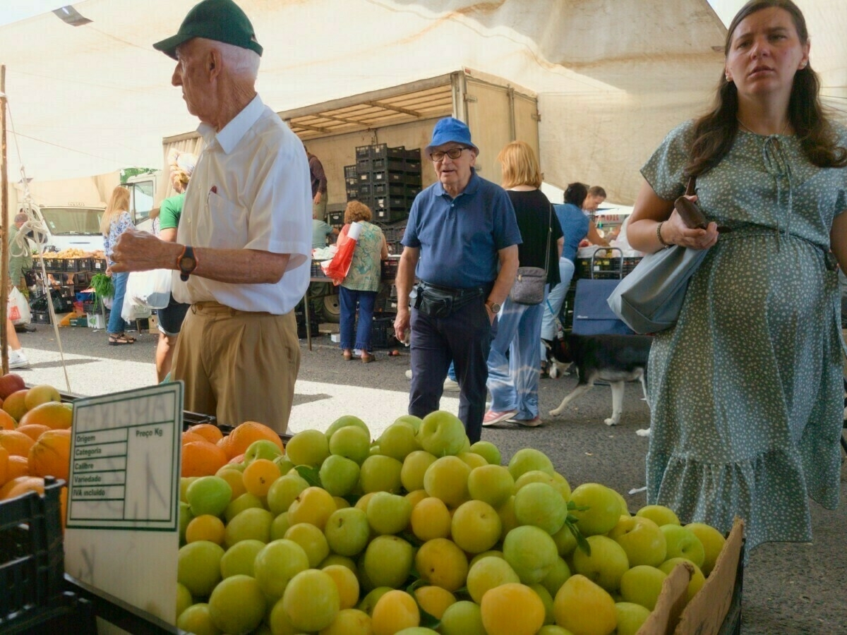 People are shopping at an outdoor market with various fruits displayed in the foreground.