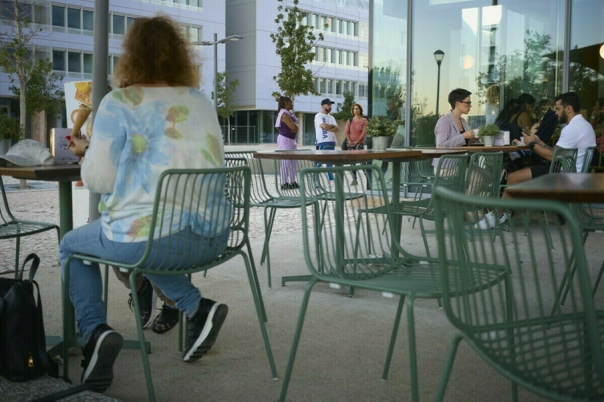 A woman sits at an outdoor café table, while other people are gathered and conversing nearby.