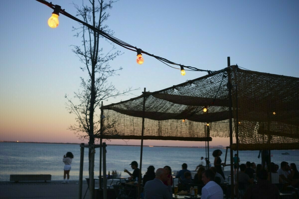 A beachfront setting at dusk features people dining under string lights and a woven canopy, with a view of the water and a pastel sky.