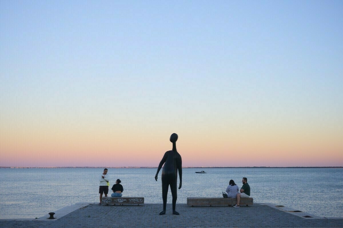 A group of people sit and stand near a tall sculpture by a calm body of water at sunset.