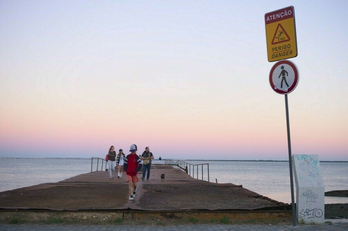 A group of people, accompanied by a dog, walks along a narrow pier that extends into a calm body of water, with warning signs in Portuguese and English visible nearby.