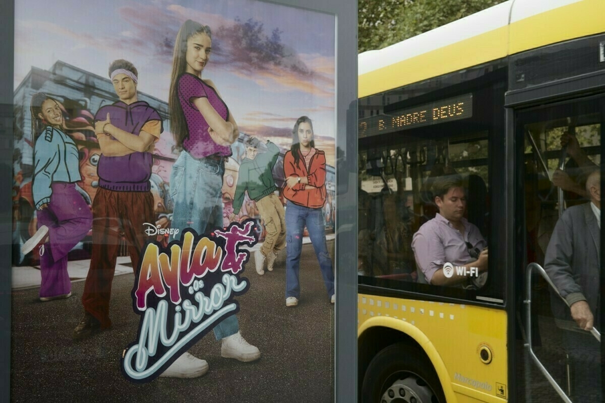 Auto-generated description: A Disney advertisement for Ayla: A Natureza de Oyá is displayed on a bus stop shelter next to a yellow city bus with passengers.