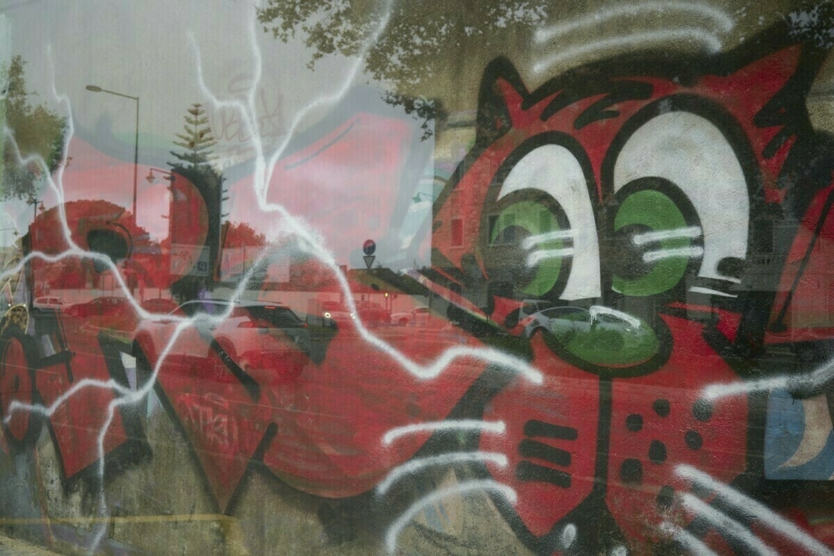 Auto-generated description: A vibrant graffiti mural featuring a cartoonish cat with green eyes and a red outline is overlaid with reflections of an urban street scene.