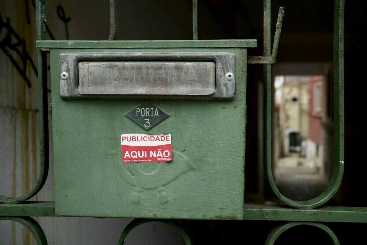 Auto-generated description: A green mailbox with a sign saying Publicidade Aqui Não (No Advertising Here) is mounted on a metal gate.