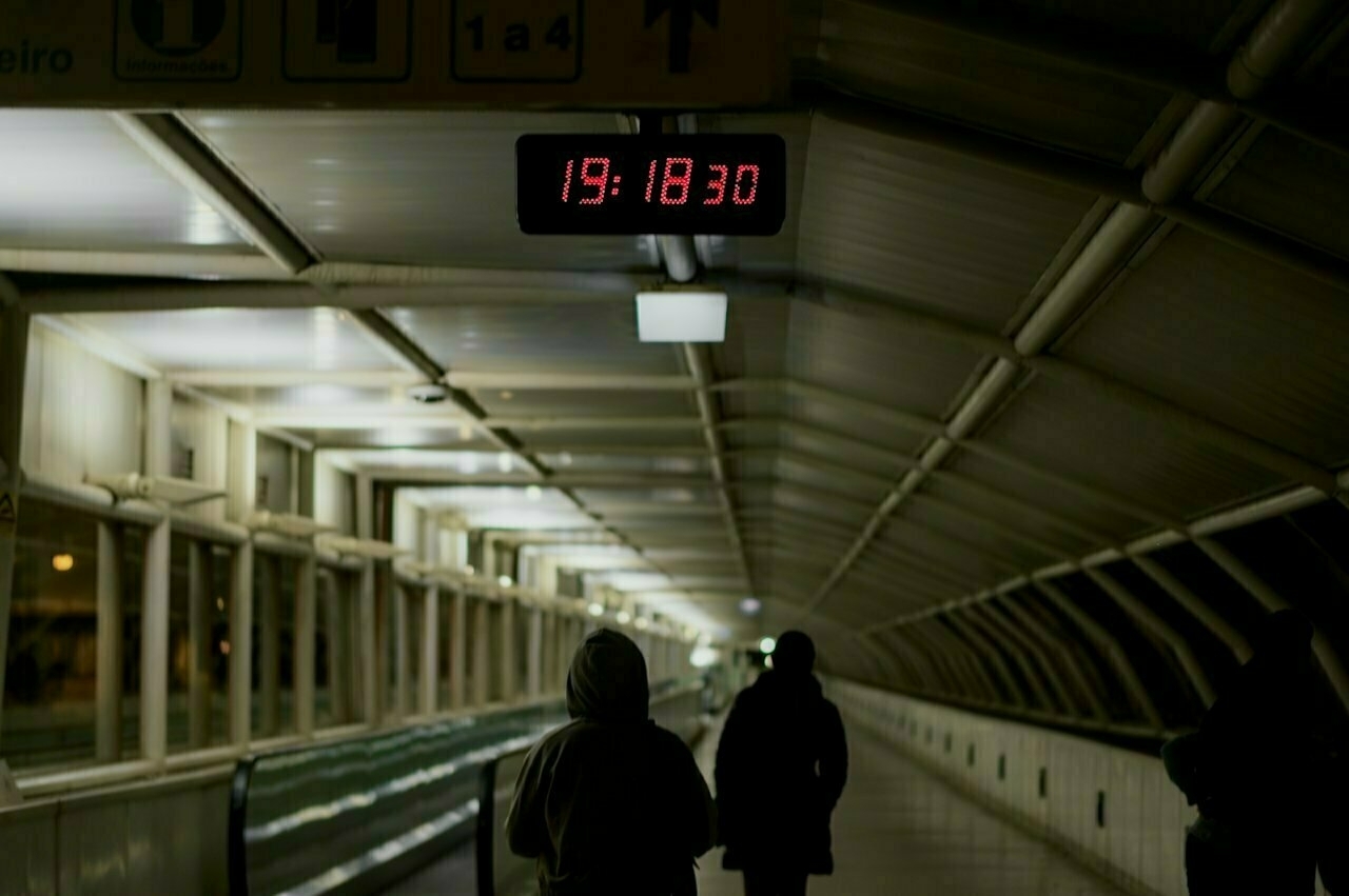 A dimly lit pedestrian tunnel features a digital clock displaying the time 19:18:30, with silhouetted figures walking beneath it.