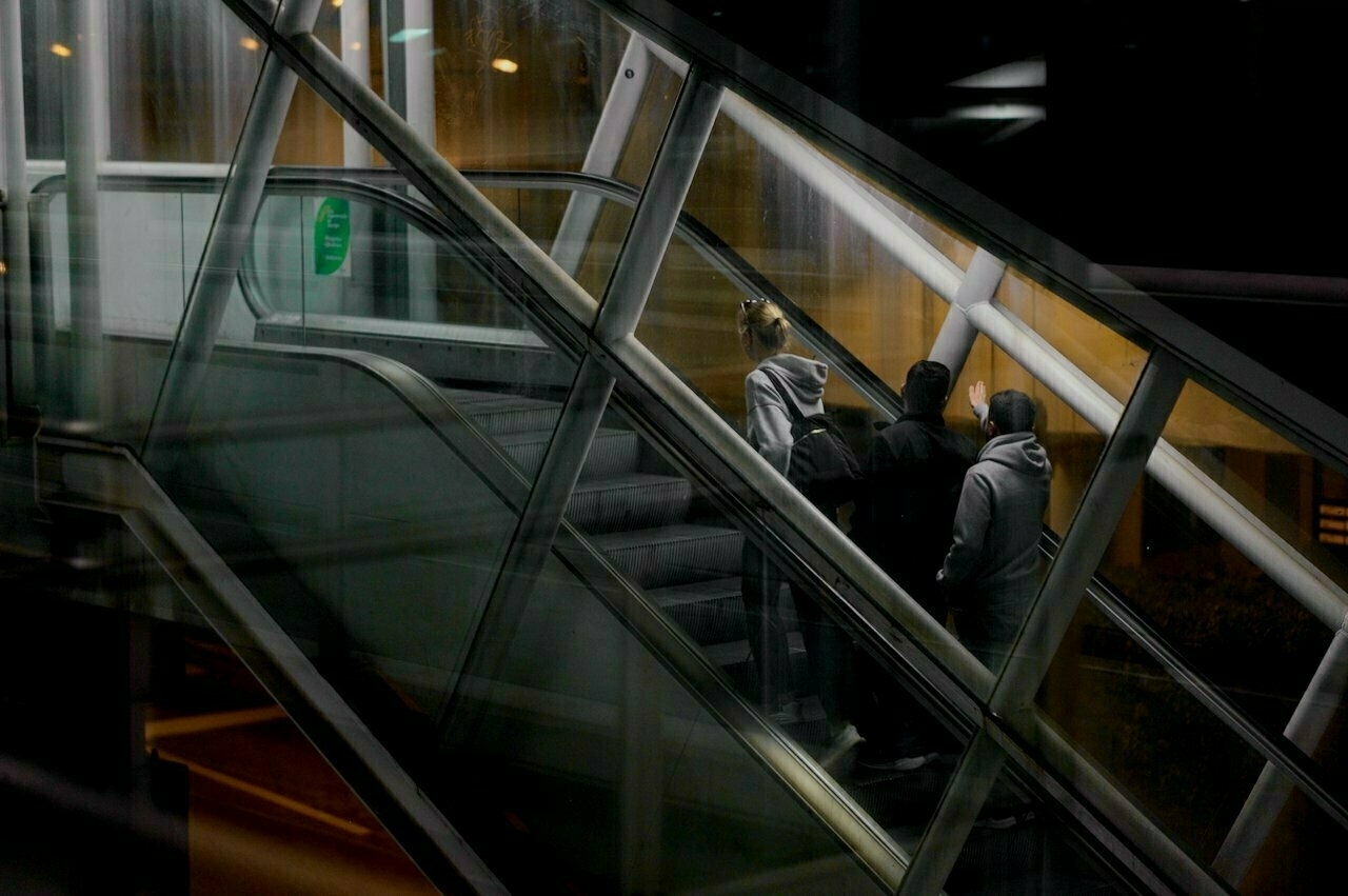 Three people wearing hoodies are ascending an escalator in a dimly lit area.