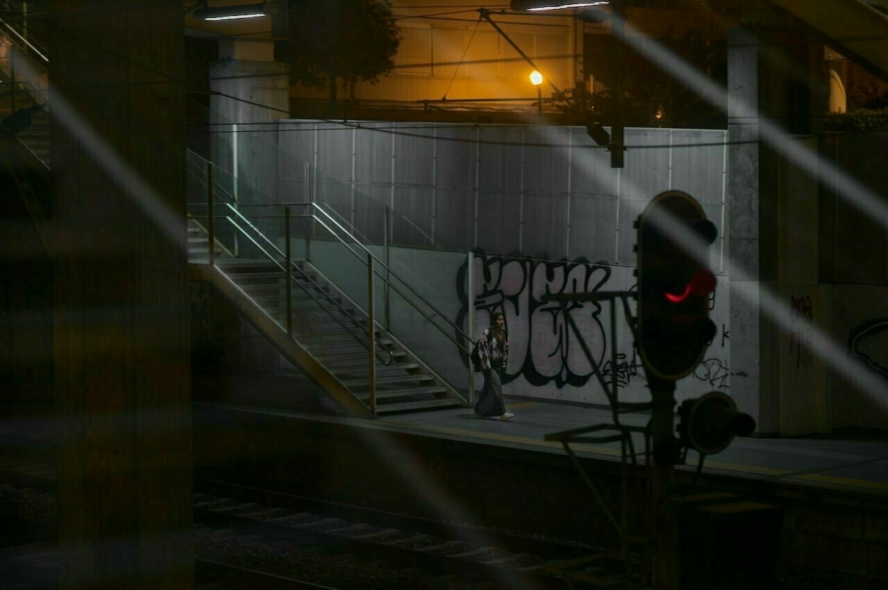 A dimly lit train station platform at night features graffiti on the wall, stairs, and a red signal light.