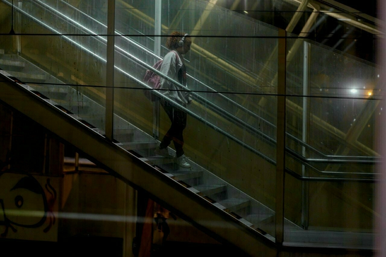 A person wearing headphones is ascending a dimly lit staircase made of glass and metal, with reflections visible on the surrounding surfaces.