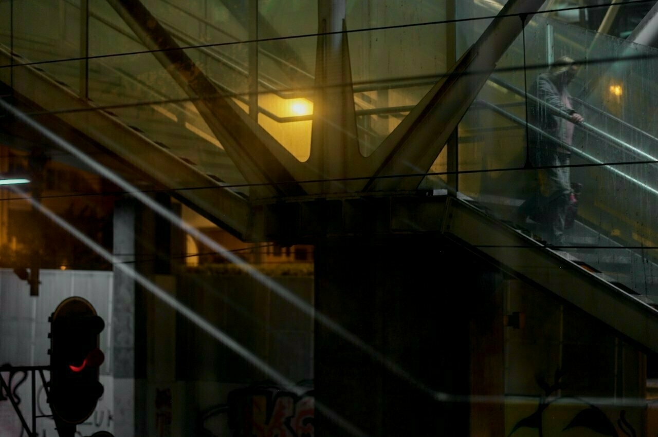 A person descends a glass-enclosed stairway at night, illuminated by nearby lights, with a red traffic signal visible in the foreground.