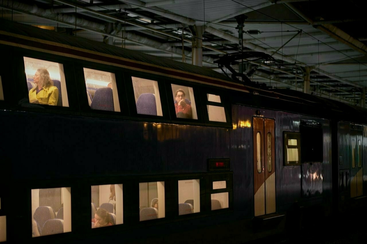 A dimly lit train at night shows passengers sitting inside, visible through the brightly lit windows.