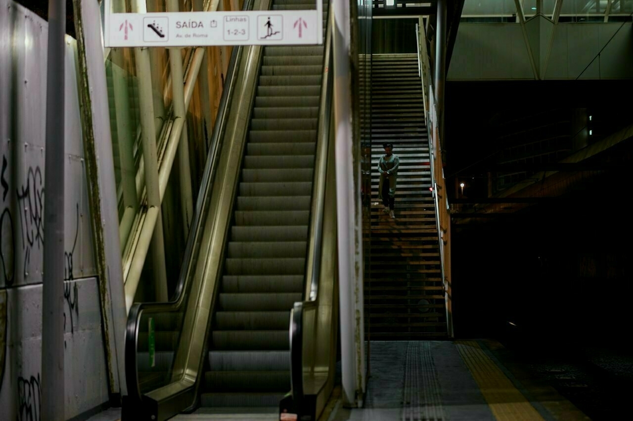 An escalator and adjacent staircase are shown in a dimly lit area with a sign indicating the way out and directions.