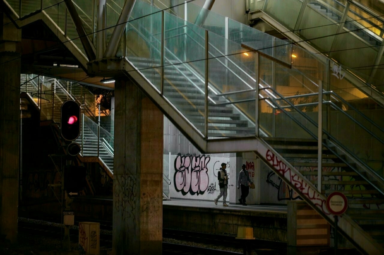 A dimly lit urban scene features staircases, graffiti, and two people walking under a bridge near a railway track.