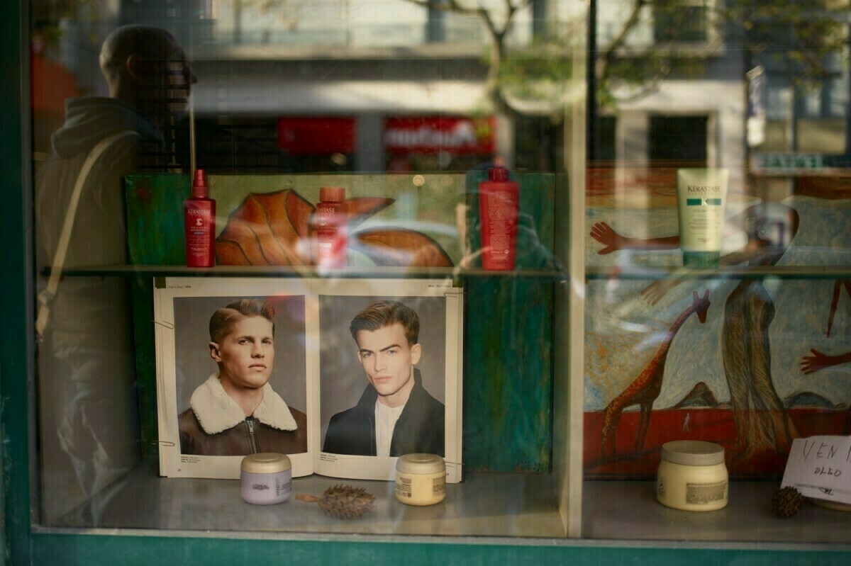 A storefront window display features grooming products and photos of two men, with artwork in the background.