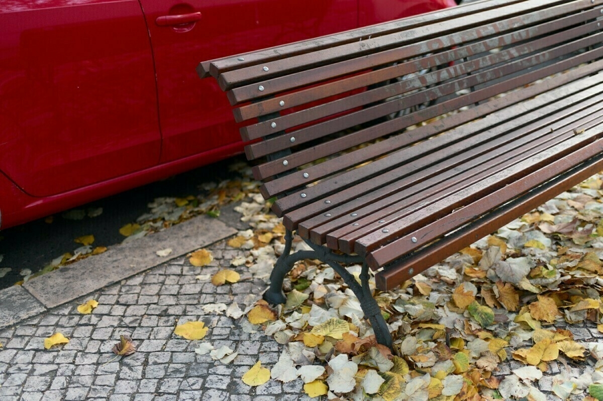 A wooden bench on a cobblestone path is surrounded by fallen leaves, with a red car parked nearby.