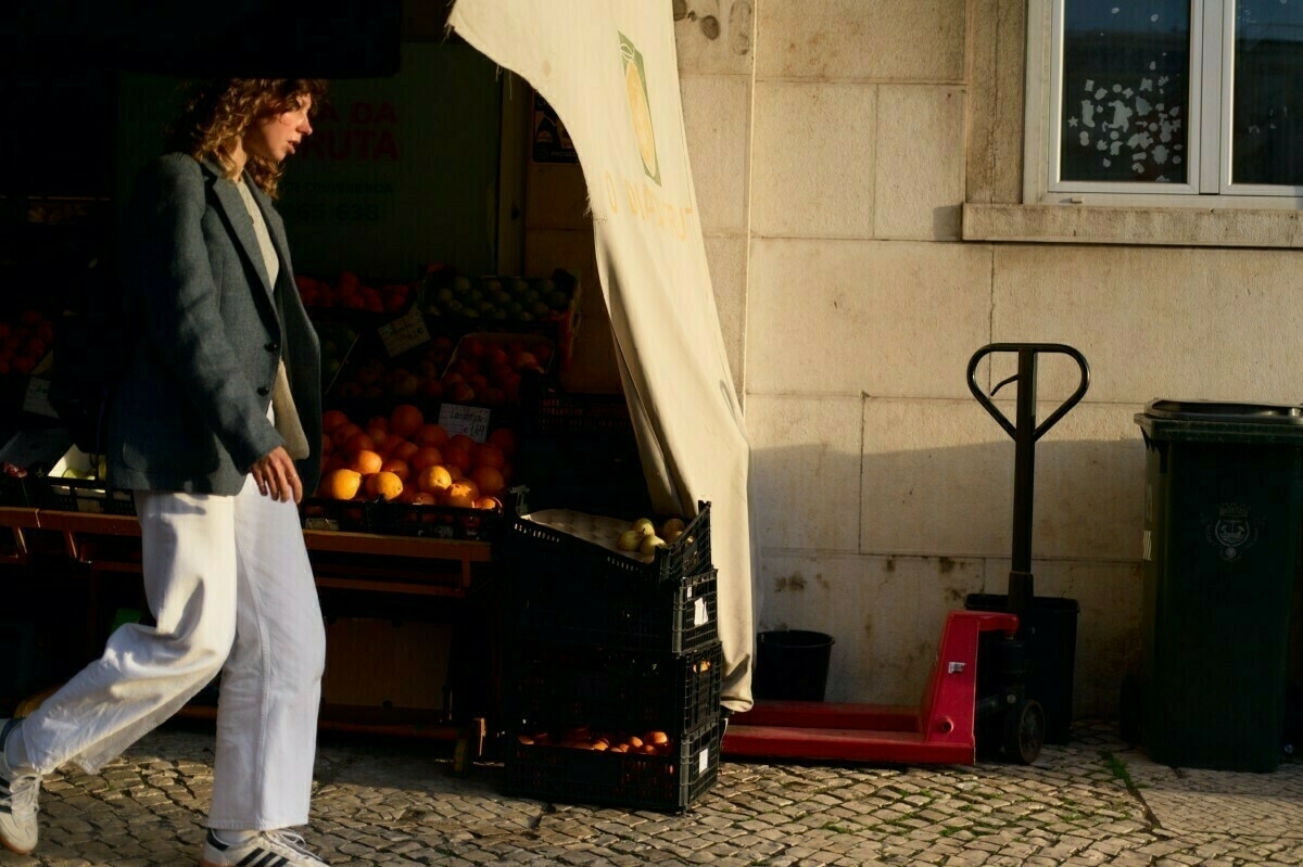 A person walks past a small fruit stand with crates of oranges and a red pallet jack on a cobblestone street.