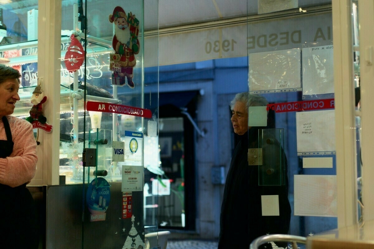 A woman stands inside a cafe decorated with holiday-themed stickers, while an older man is visible outside through the glass door.