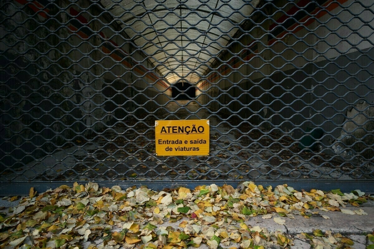 A gated tunnel with a warning sign in Portuguese and fallen leaves scattered on the ground.