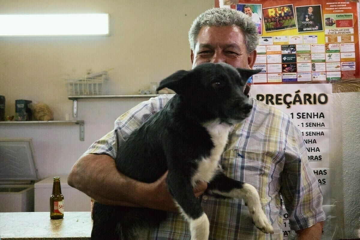 A man holding a black and white dog stands in a room with a variety of posters and a beer bottle on the table.