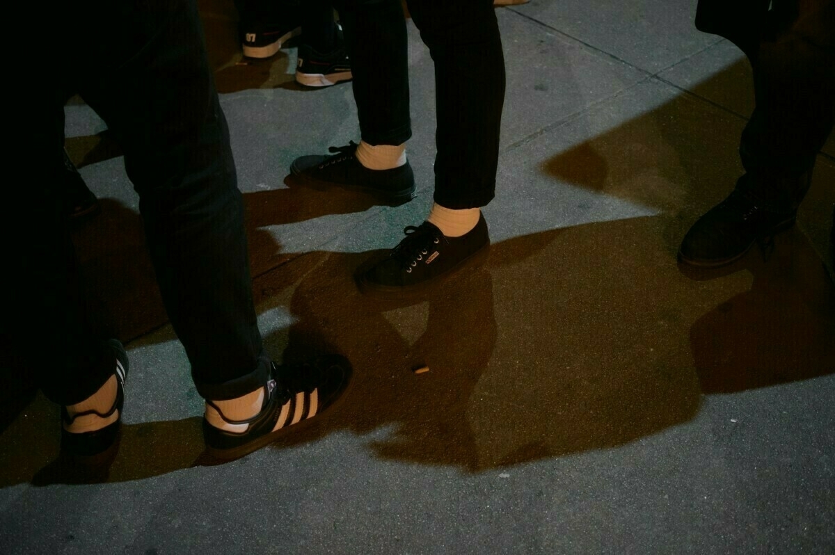 People are standing on a dimly lit sidewalk, showing only their legs and feet wearing sneakers.