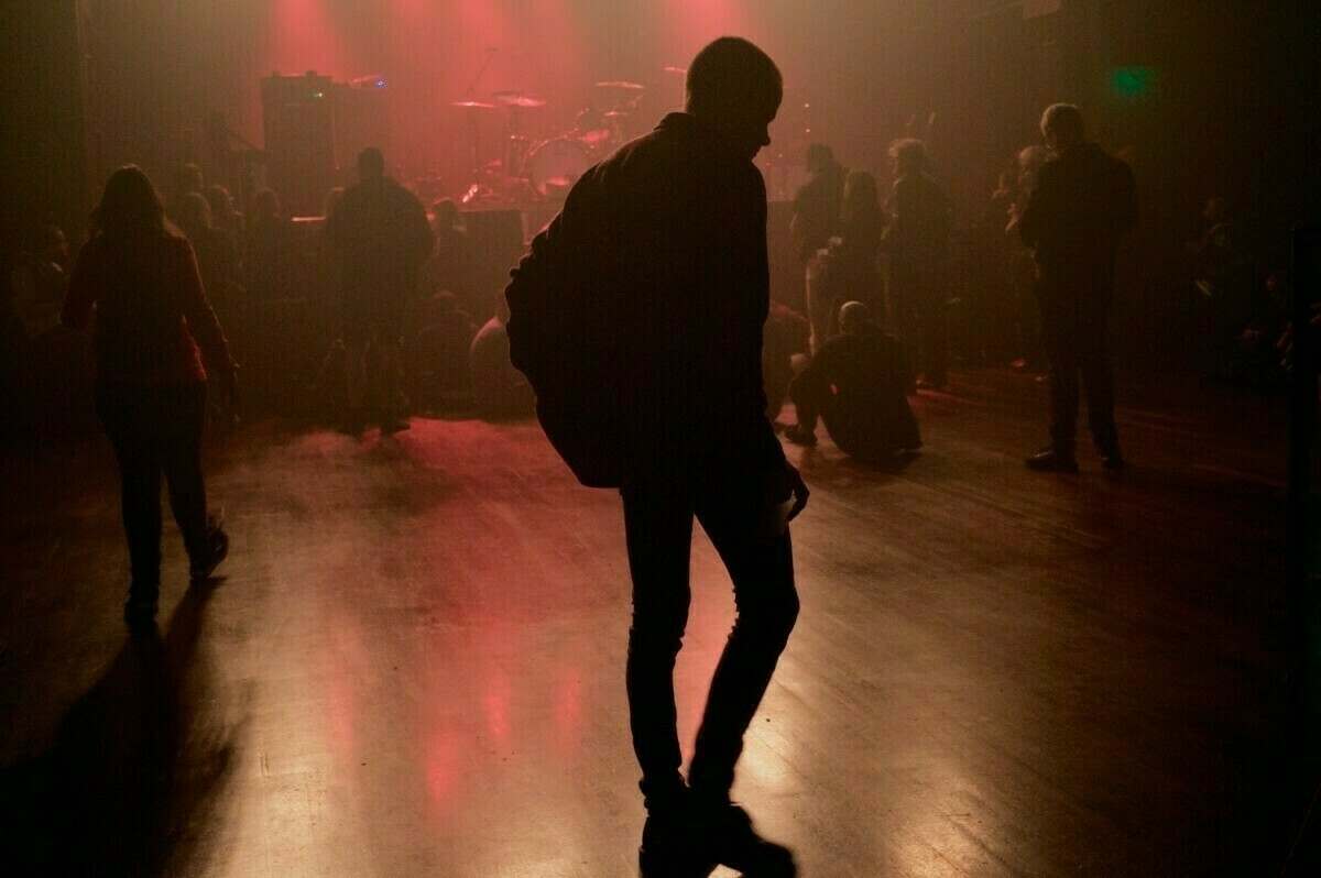 A silhouette of a person stands in a dimly lit venue with a small crowd and red stage lights in the background.