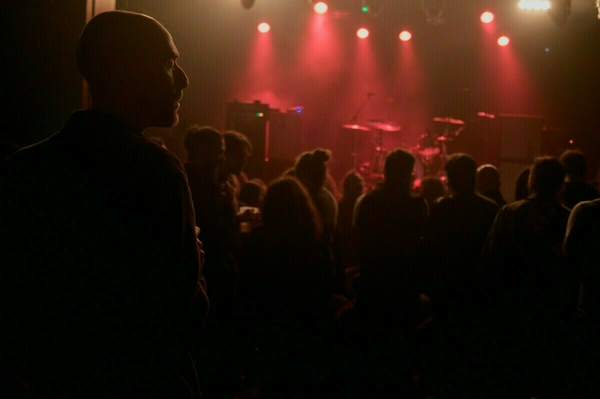 A crowd of people is gathered in a dimly lit concert venue with red stage lights illuminating the area.