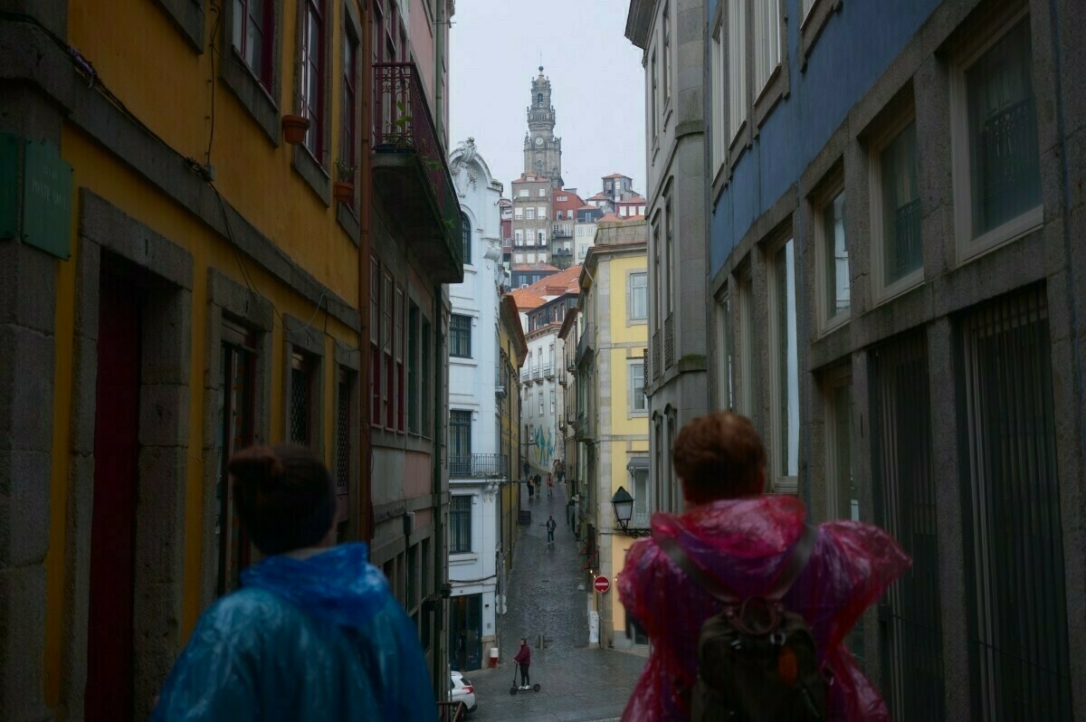 A narrow street lined with colorful buildings leads to a distant Clérigos Tower, with two people in rain ponchos walking down the street.