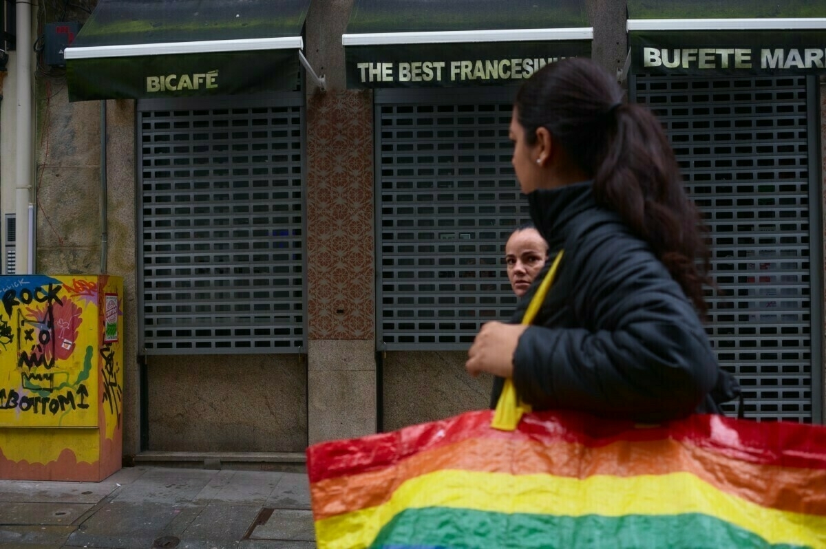 A person carrying a large rainbow-colored bag walks in front of closed storefronts, with graffiti on a nearby utility box.