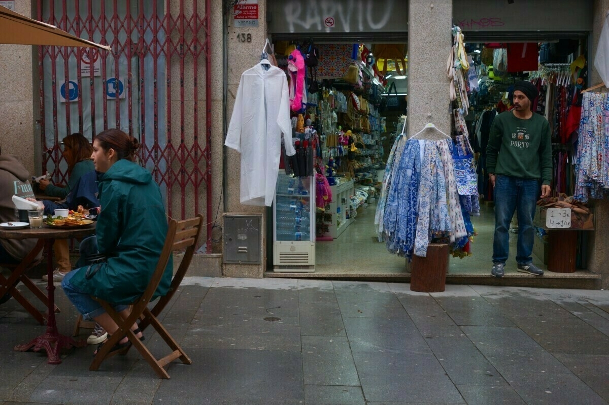 Two people are sitting at an outdoor table near a shop with clothes displayed at the entrance, while a person stands in the shop doorway.