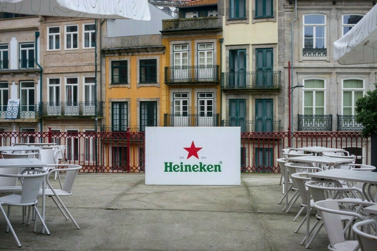 A café terrace with empty white tables and chairs is set against a backdrop of colorful, historic buildings, featuring a prominent Heineken sign.