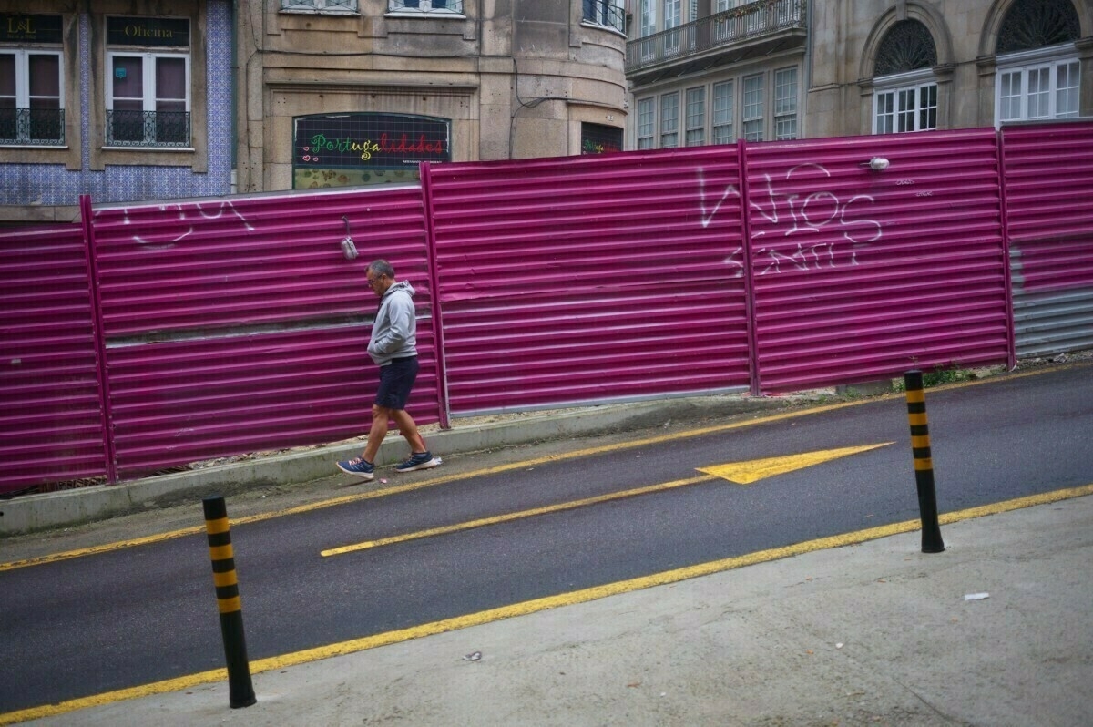 A person walks along a street lined with a tall, pink fence in front of a building.