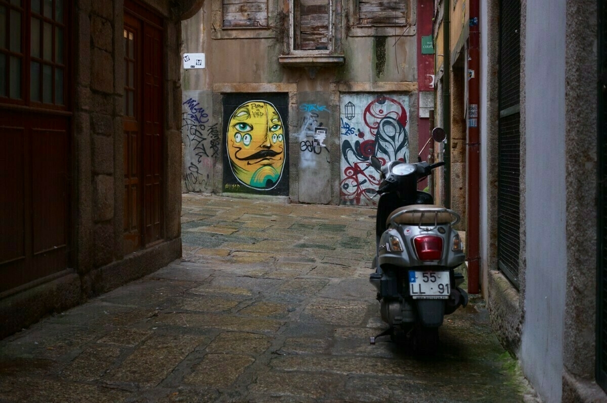 A narrow alley features a scooter parked on cobblestone pavement, with colorful graffiti, including a yellow face, on the walls.