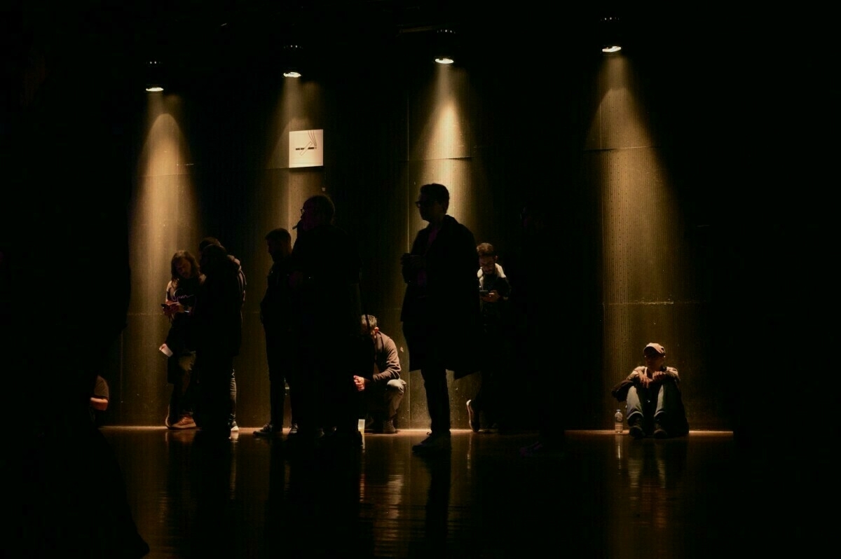 People are silhouetted against a dark background, standing and sitting under spotlights in a dimly lit area.