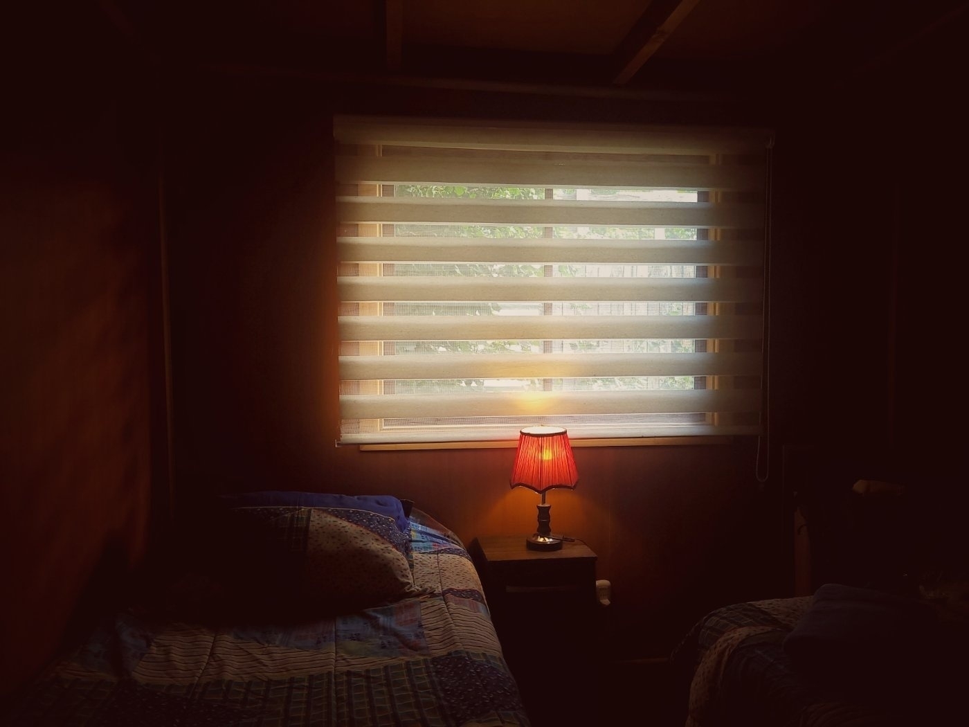 A dimly lit room features two beds and a warmly lit lamp on a small table in front of a window with horizontal blinds.