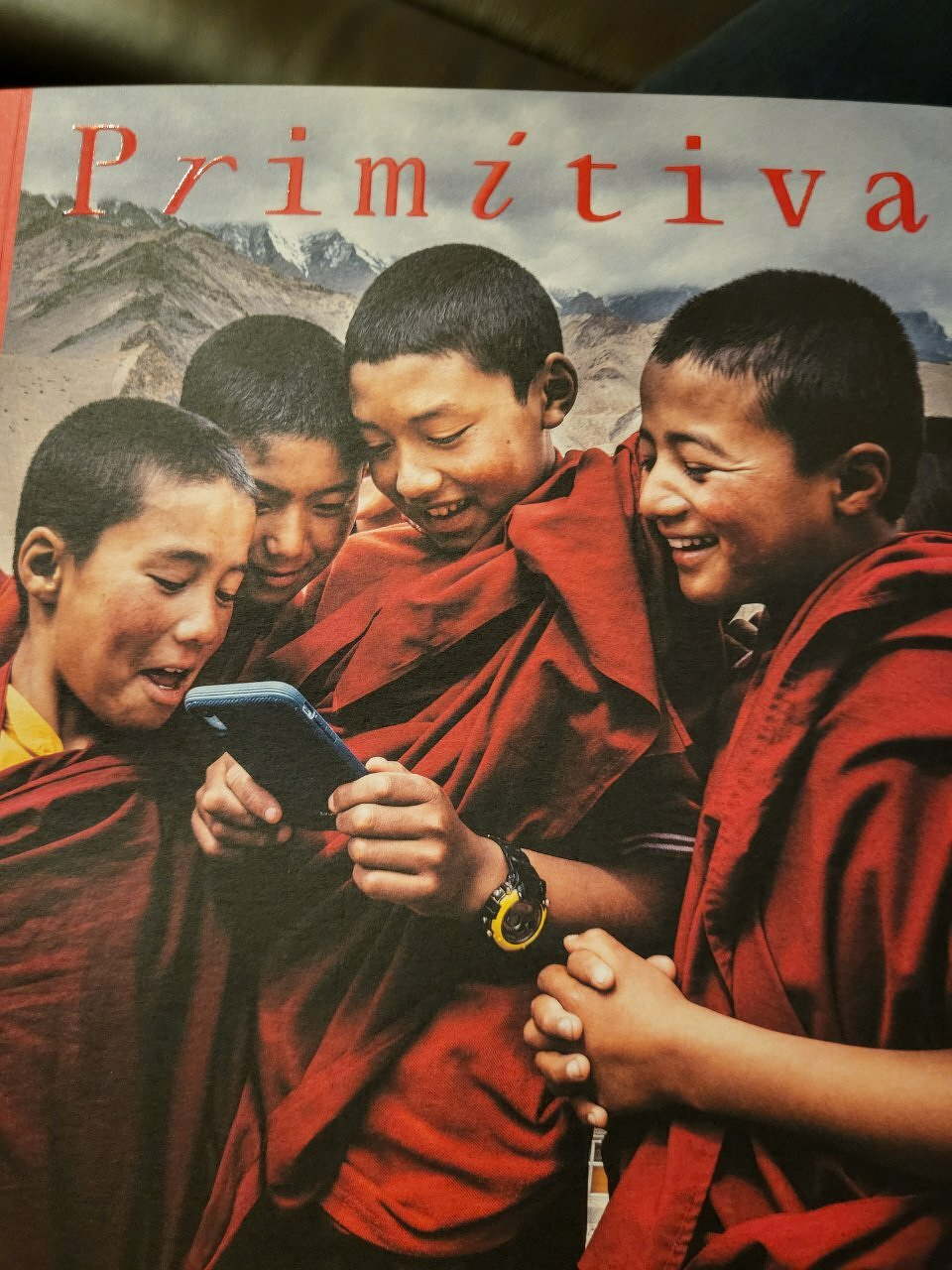 Four boys dressed in red robes are joyfully gathered around a smartphone while standing outdoors.