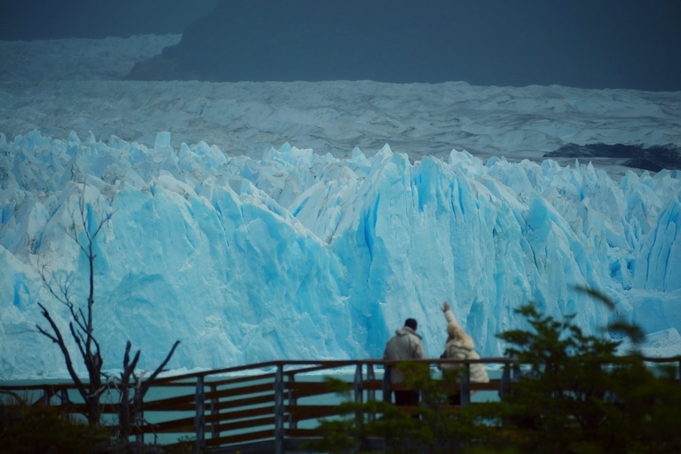 Two people stand on a wooden platform admiring a vast, icy glacier under a moody sky.