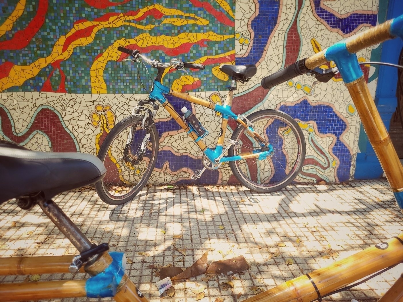 A blue bicycle and a bamboo-framed bike are parked in front of a colorful mosaic wall with abstract designs.