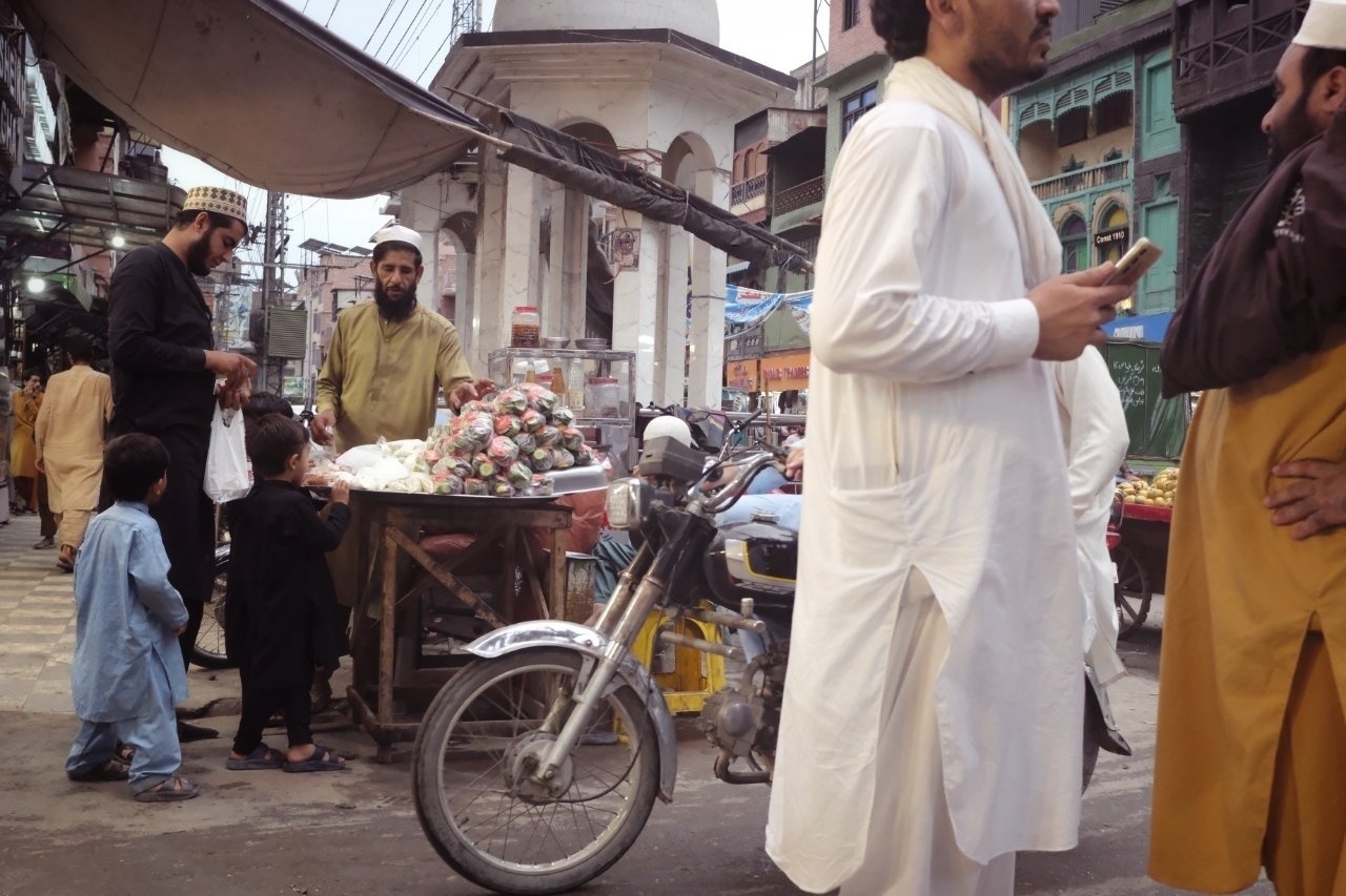 A busy street scene features people chatting near a fruit stall, a motorcycle, and various shops in the background.