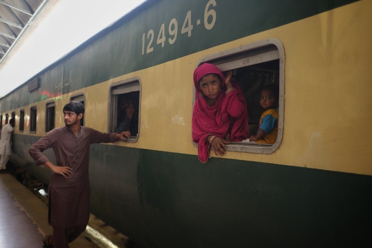 A person in pink, along with a child, looks out of a train window while another person stands on the platform beside the train.