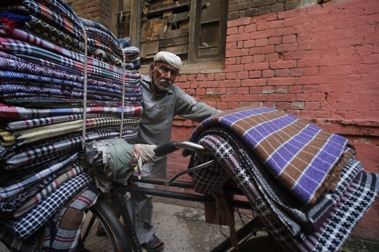 A man in traditional attire pushes a cycle rickshaw loaded with stacks of colorful textiles along a brick-lined street.