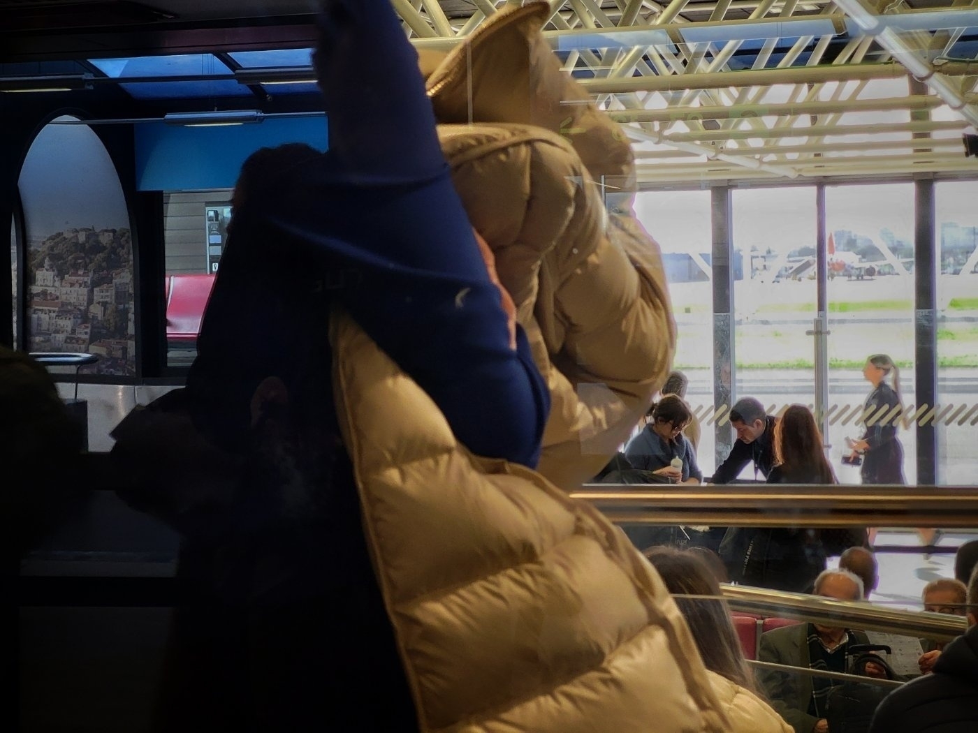 A person in a beige puffy jacket is reflected in a glass surface at an indoor terminal with people and large windows in the background.