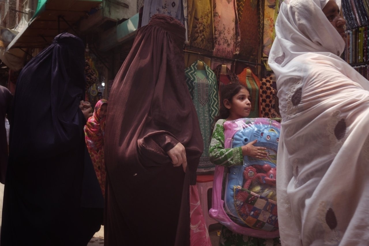 A young girl carrying a colorful backpack stands among women wearing burkha on a bustling street.