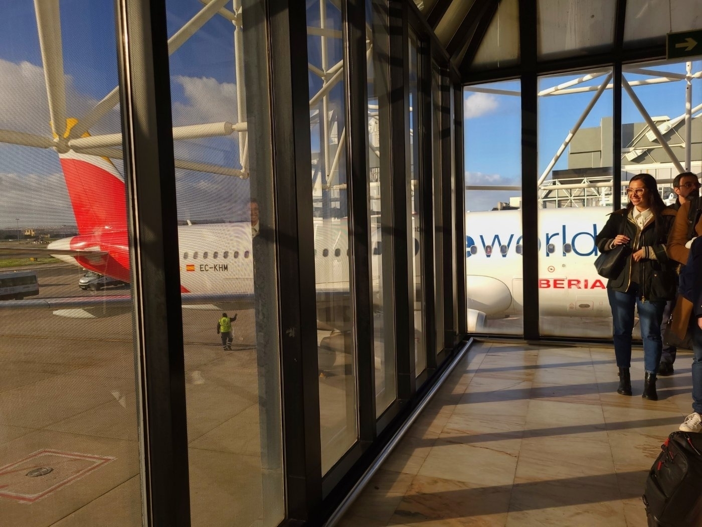 People walk through an airport terminal with an Iberia airplane visible outside the windows.