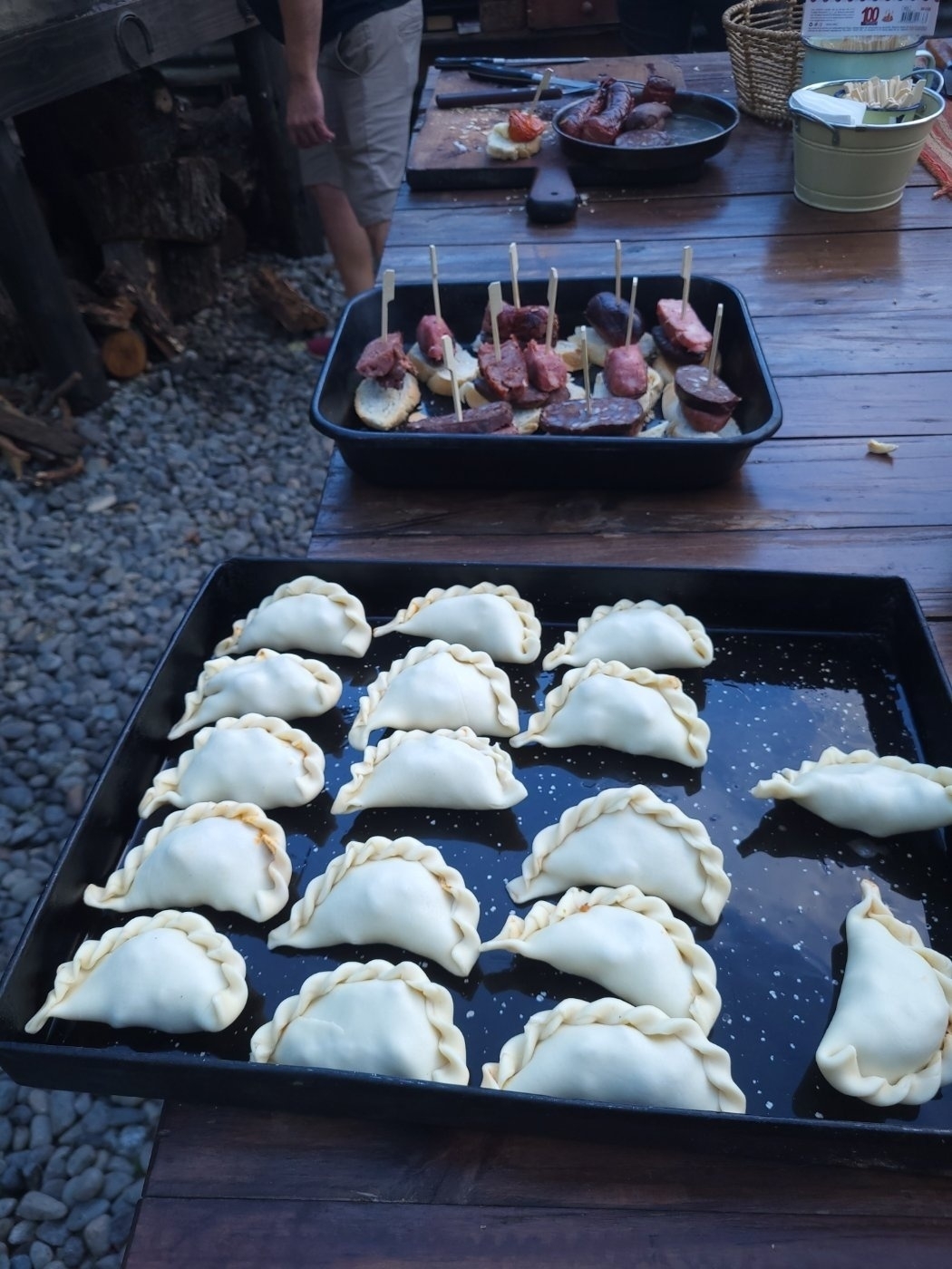 Trays of uncooked empanadas and skewered appetizers are displayed on a wooden table outdoors.