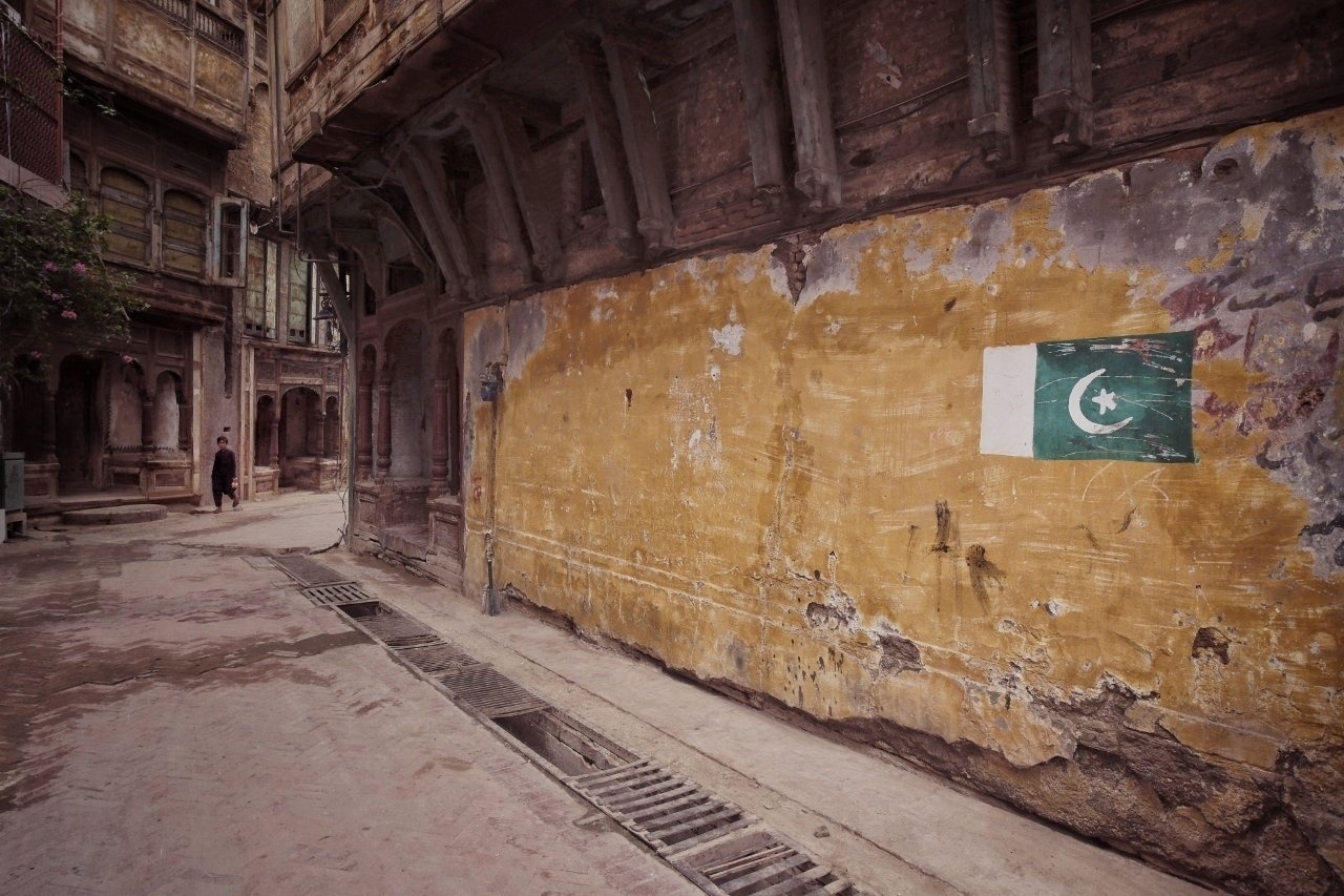 A narrow, worn street with a yellowed wall displaying the Pakistani flag, lined with aged architecture, leads to a person in the distance.
